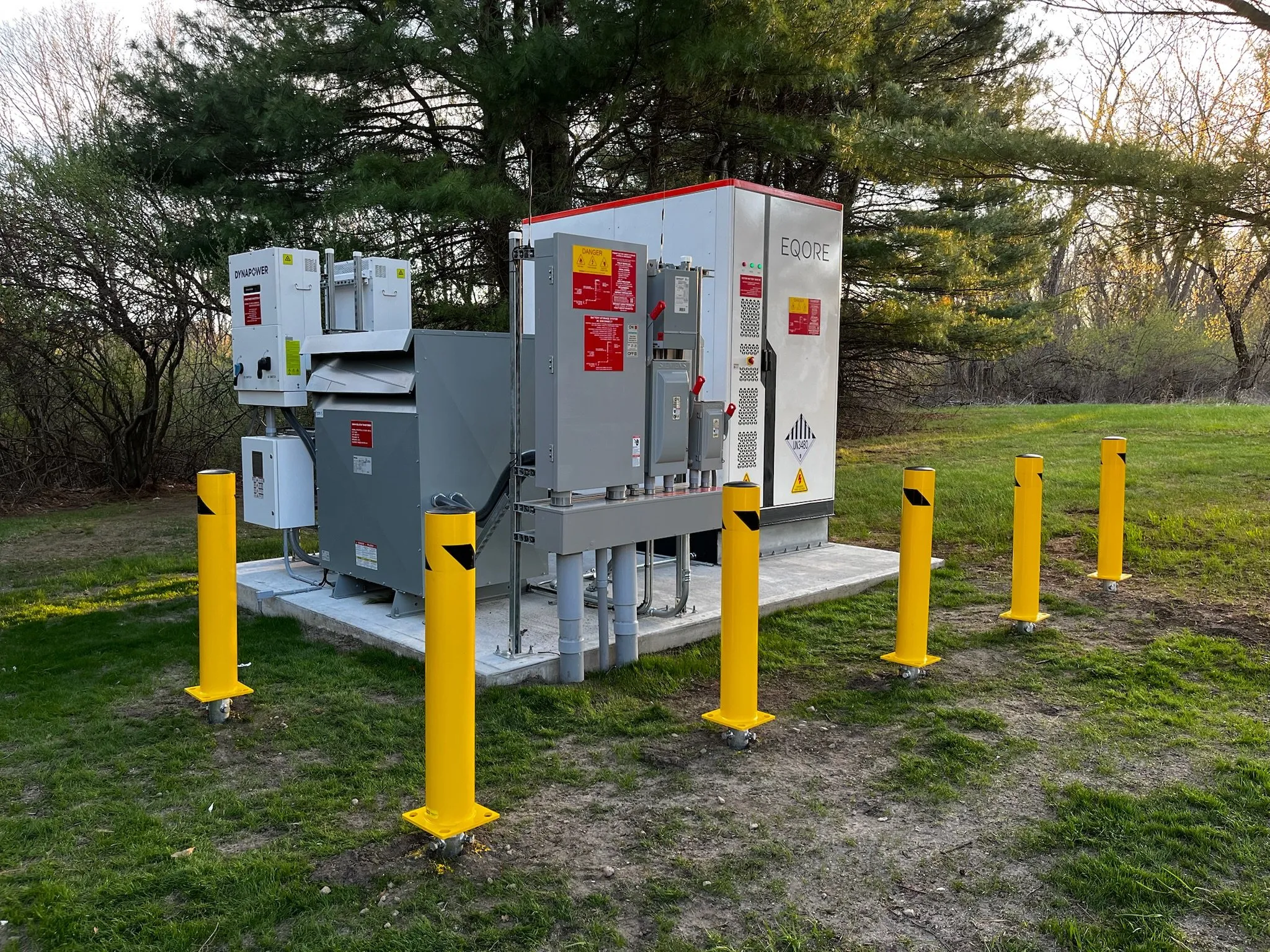 Outdoor battery storage system on a concrete pad, surrounded by yellow protective bollards.